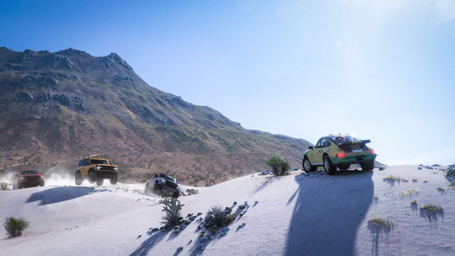 Four off-road vehicles racing over snow-covered hills beneath a clear bright blue sky with mountains in the background