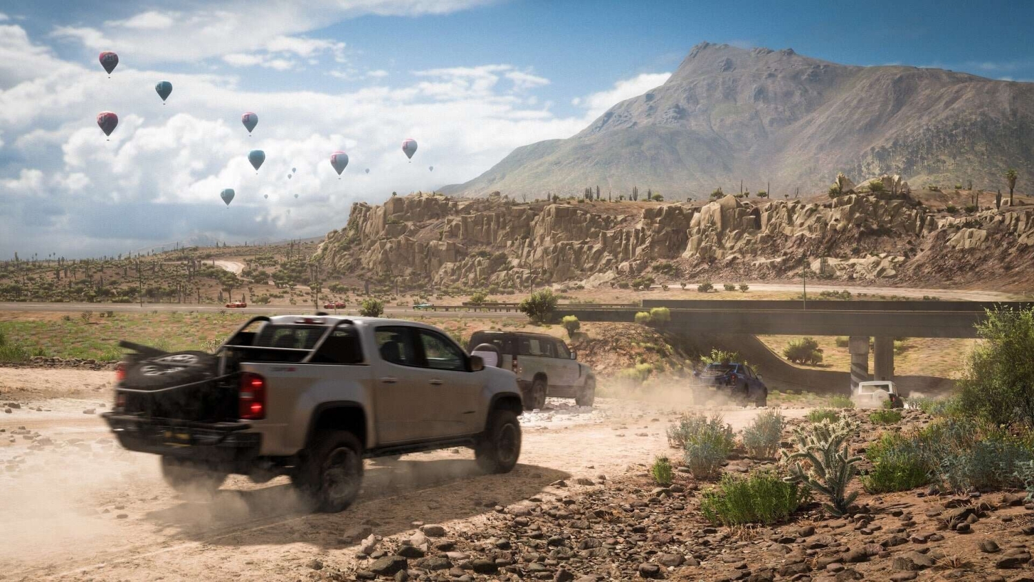 Four off-road vehicles driving on a dusty desert track with hot air balloons floating in the sky