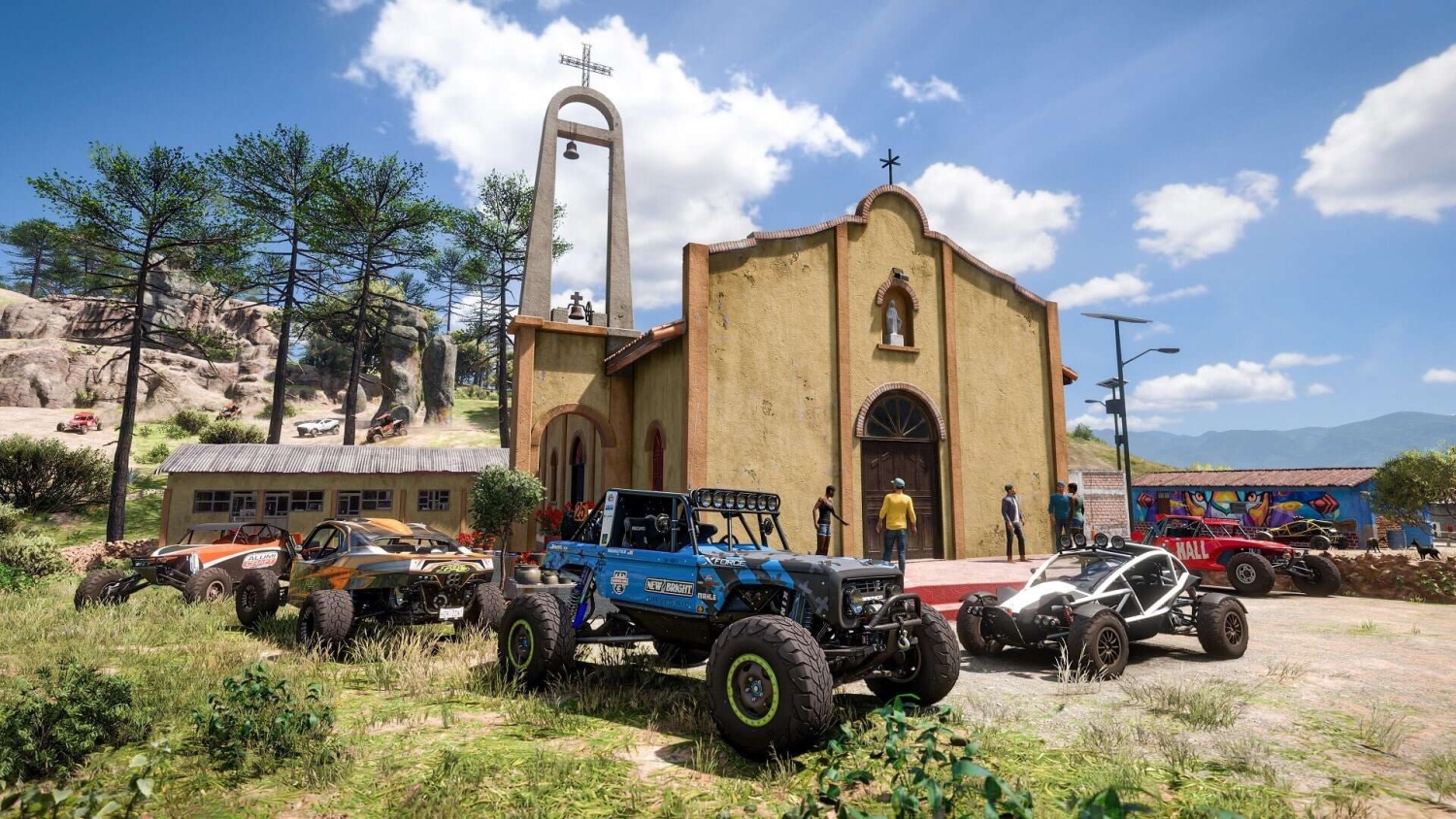 Custom off-road vehicles parked beside a small rustic yellow church with bell tower under a bright blue sky