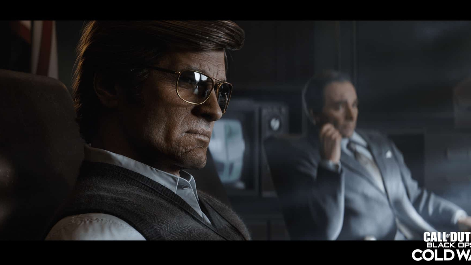 Two men seated in a dark room, one wearing glasses and a sweater vest, the other in a suit, with a vintage TV in the background