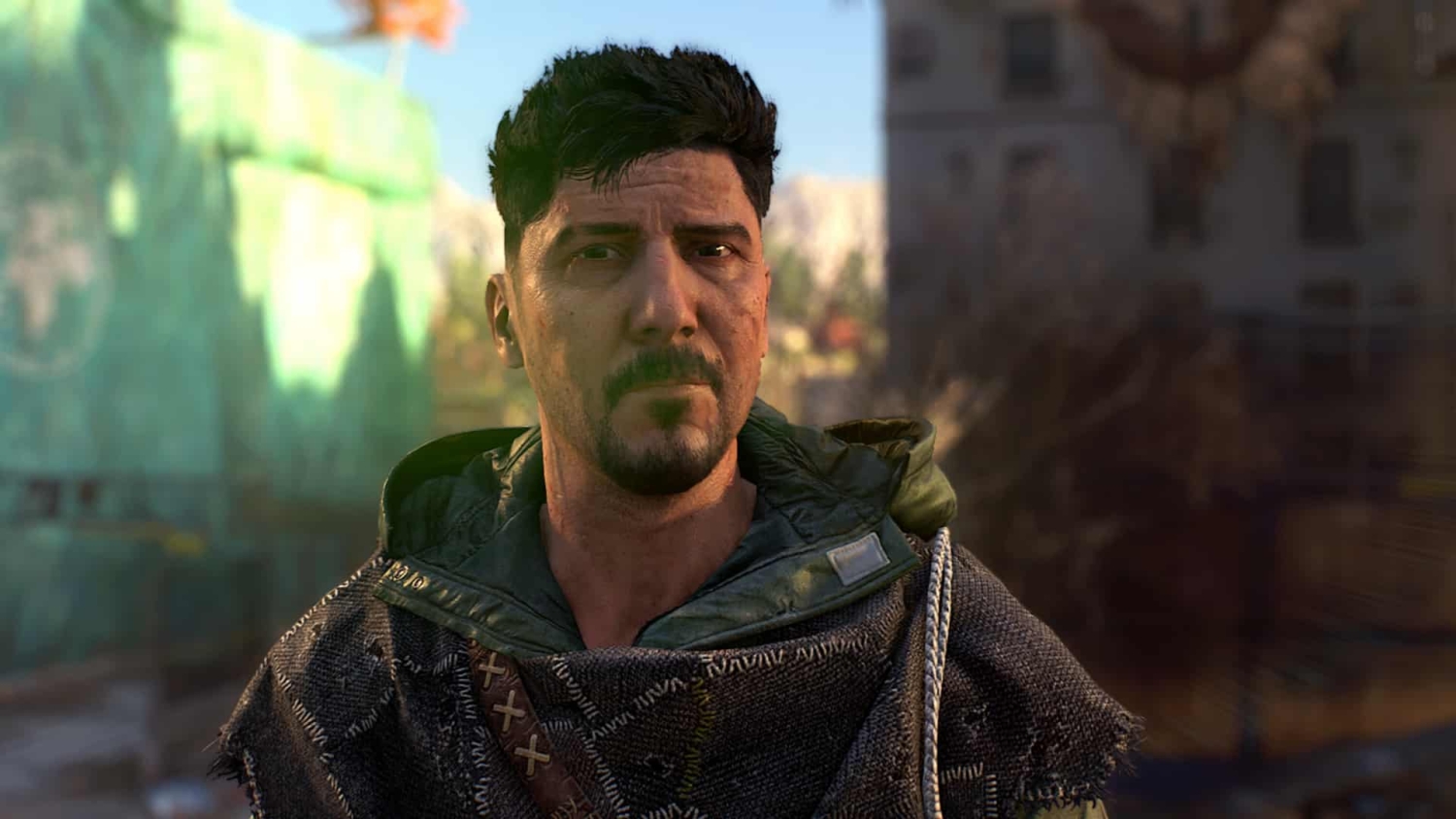 Close-up of a man with dark hair and stubble wearing patched clothing and a hood in a blurred urban setting