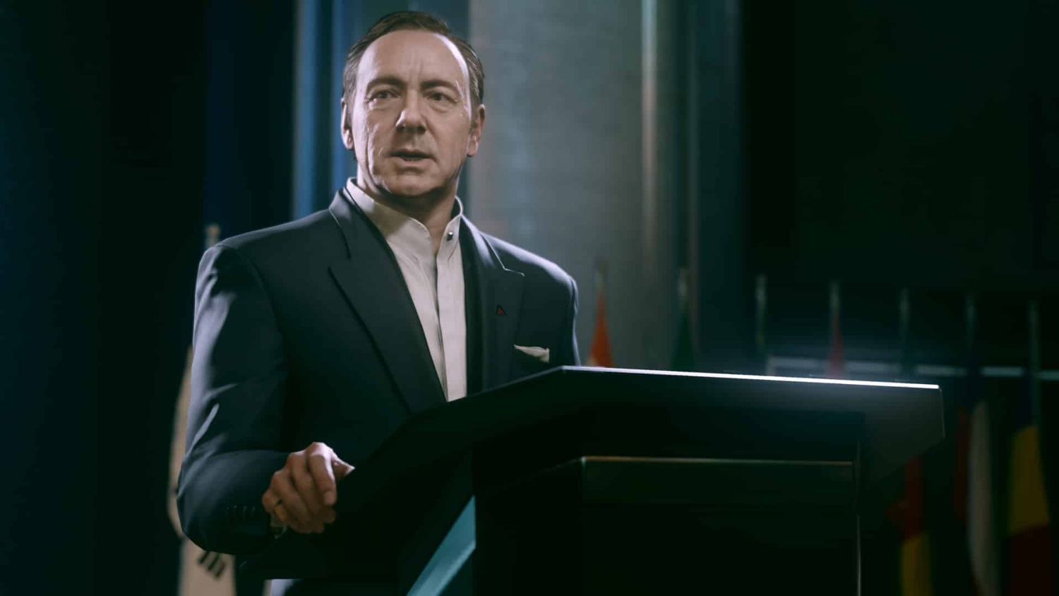 Man in dark suit and white shirt speaking at a podium in a dimly lit hall with flags behind him