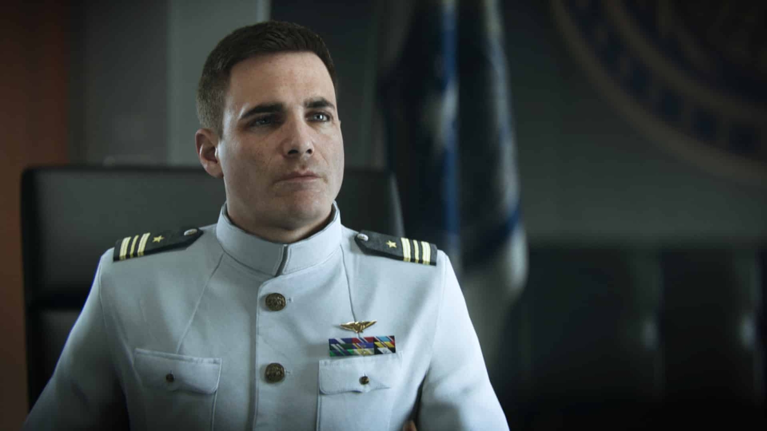 Man in white naval uniform with medals sitting in an office with a dark background