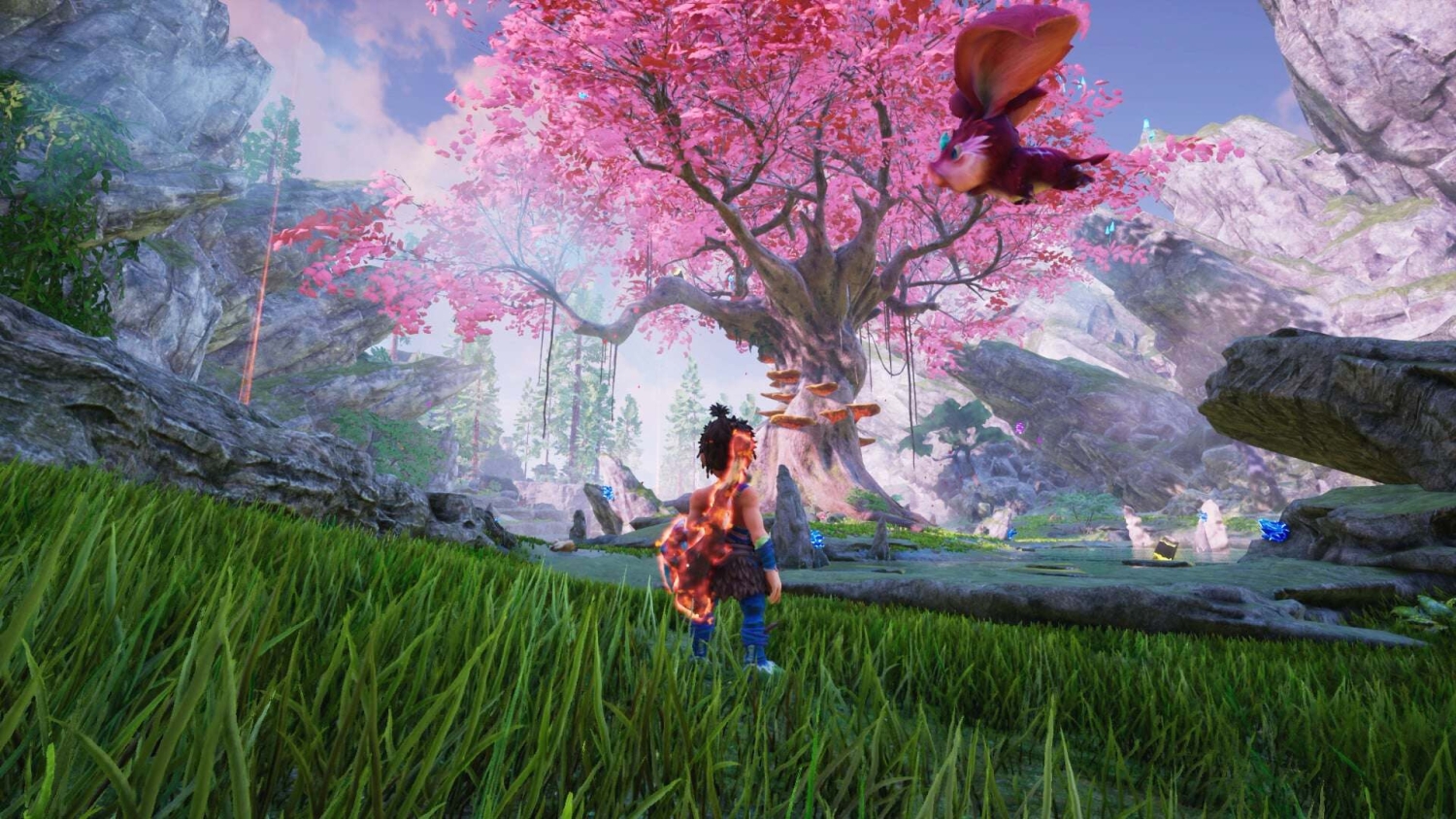 A character with fiery aura stands on green grass looking at a large pink blossomed tree in a rocky valley