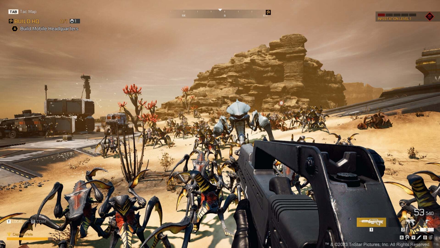 First-person shooter view targeting a swarm of alien creatures in a desert with rocky terrain and futuristic structures