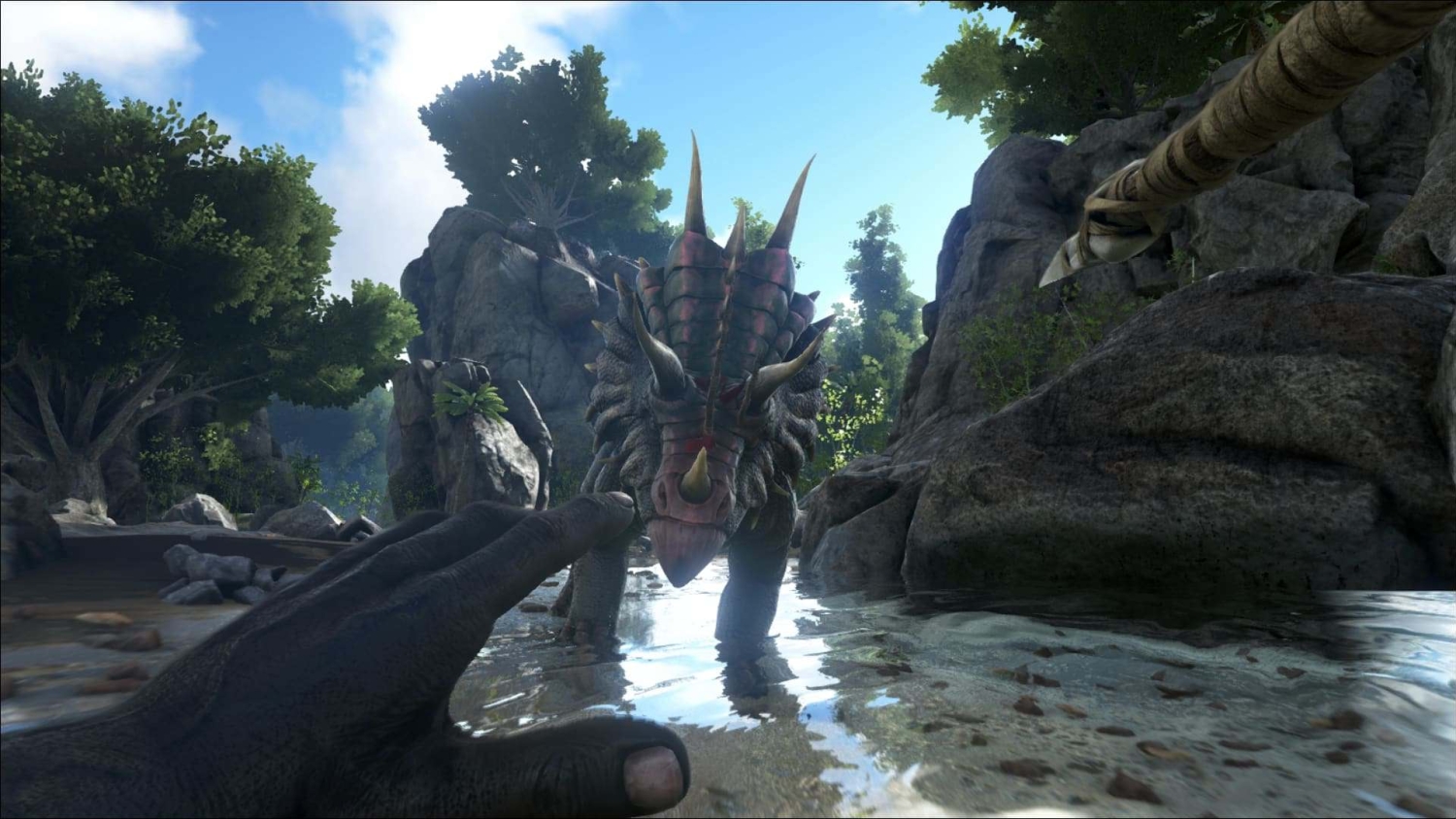 First-person view of a hand reaching towards a horned dinosaur standing in shallow river water surrounded by rocks and trees