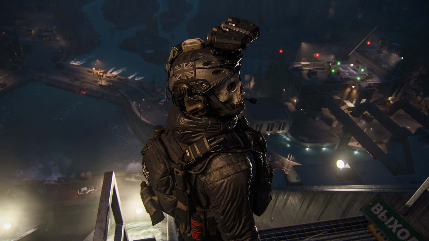 Soldier wearing tactical gear and skull mask stands on rooftop overlooking a lit military base at night