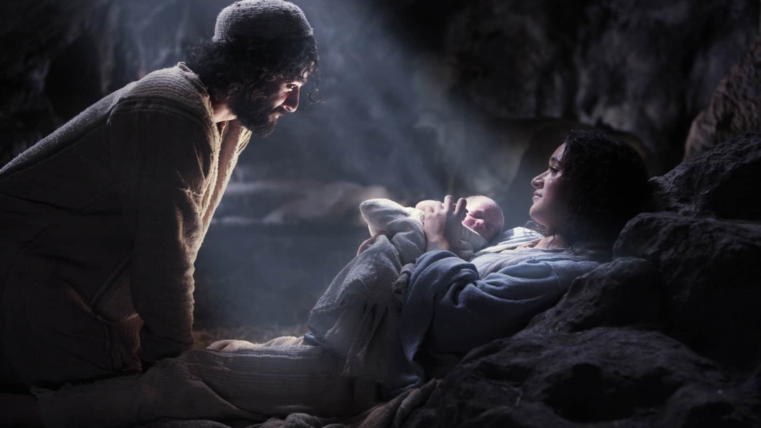 Man and woman in robes beside newborn baby lying in cave lit by soft beam of light
