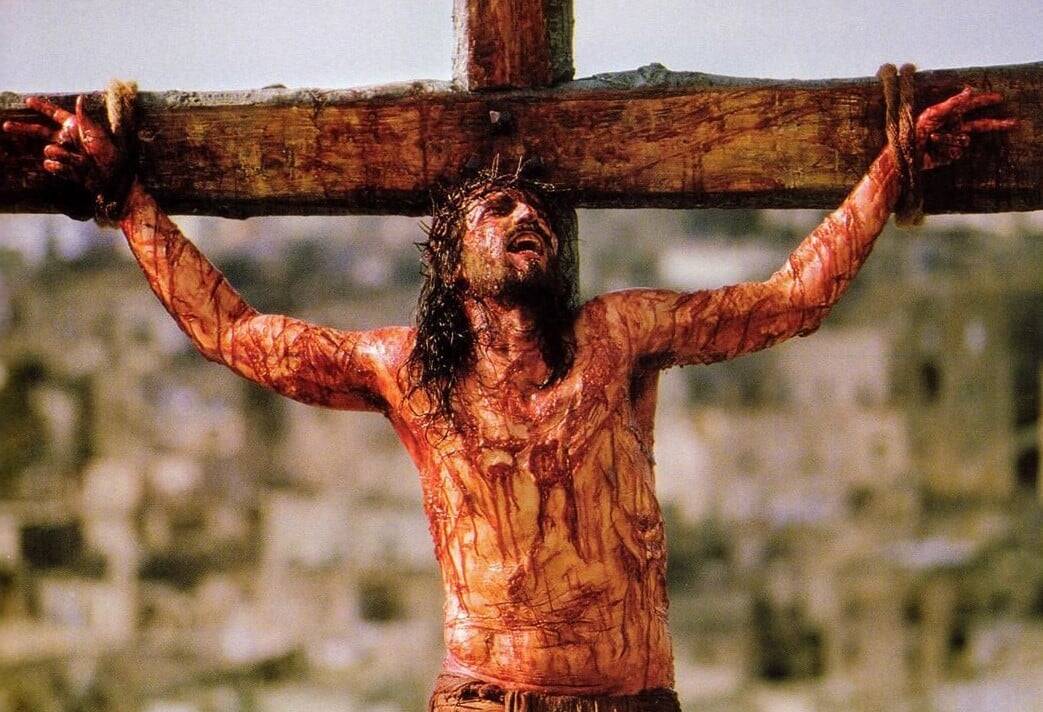 Man bound on a wooden cross, covered in blood and wounds, looking upwards in agony