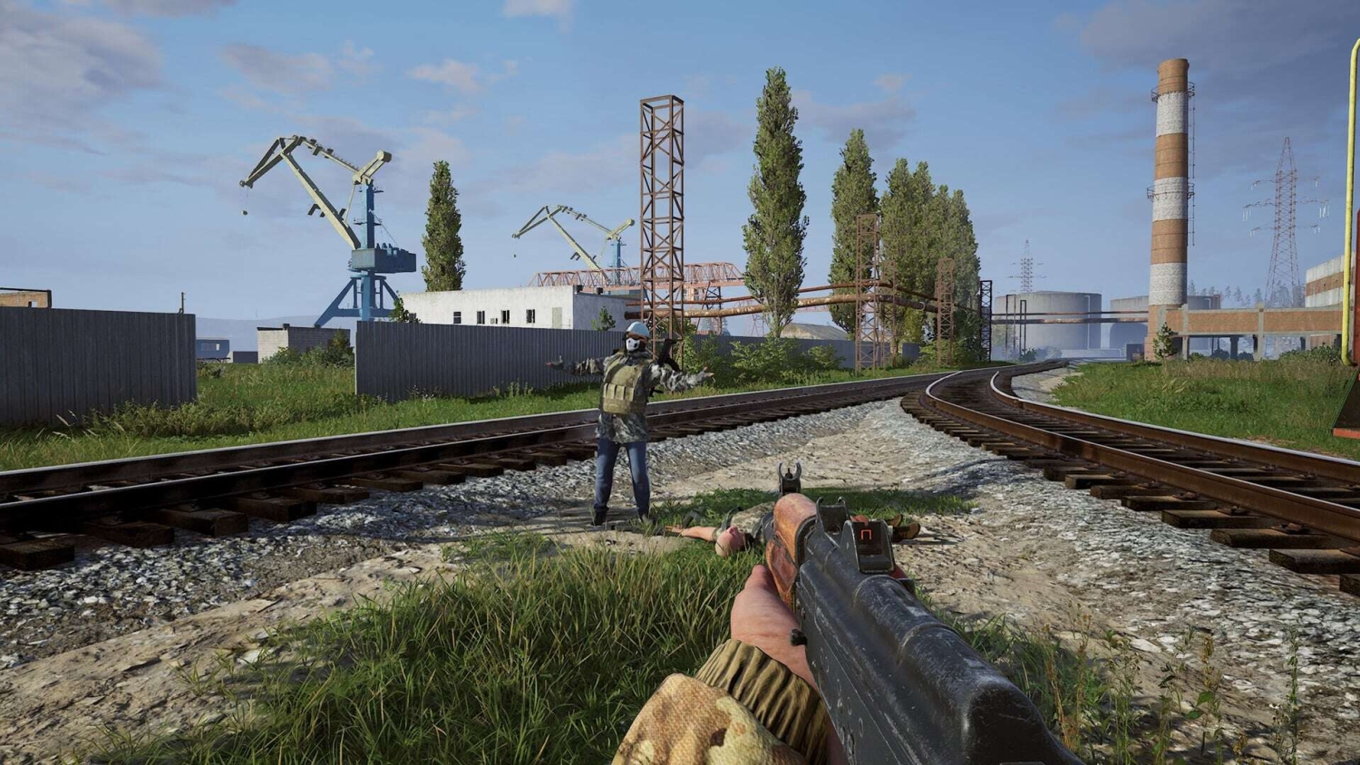 First-person shooter view aiming at an armed figure standing on railway tracks in an industrial environment with cranes and pipelines