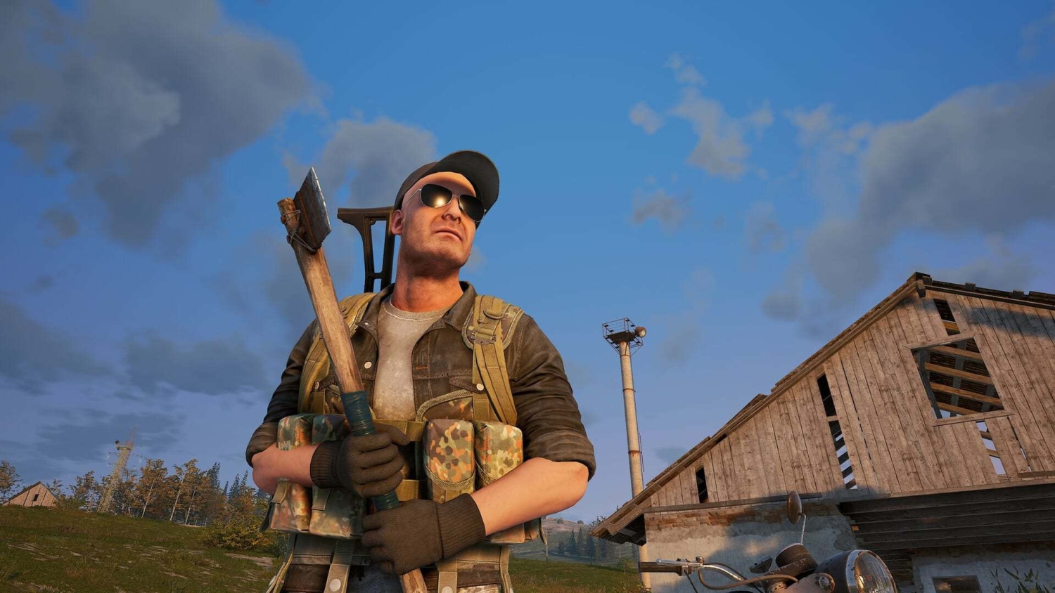Man wearing sunglasses and cap holding an axe in front of a rustic barn under blue sky with clouds