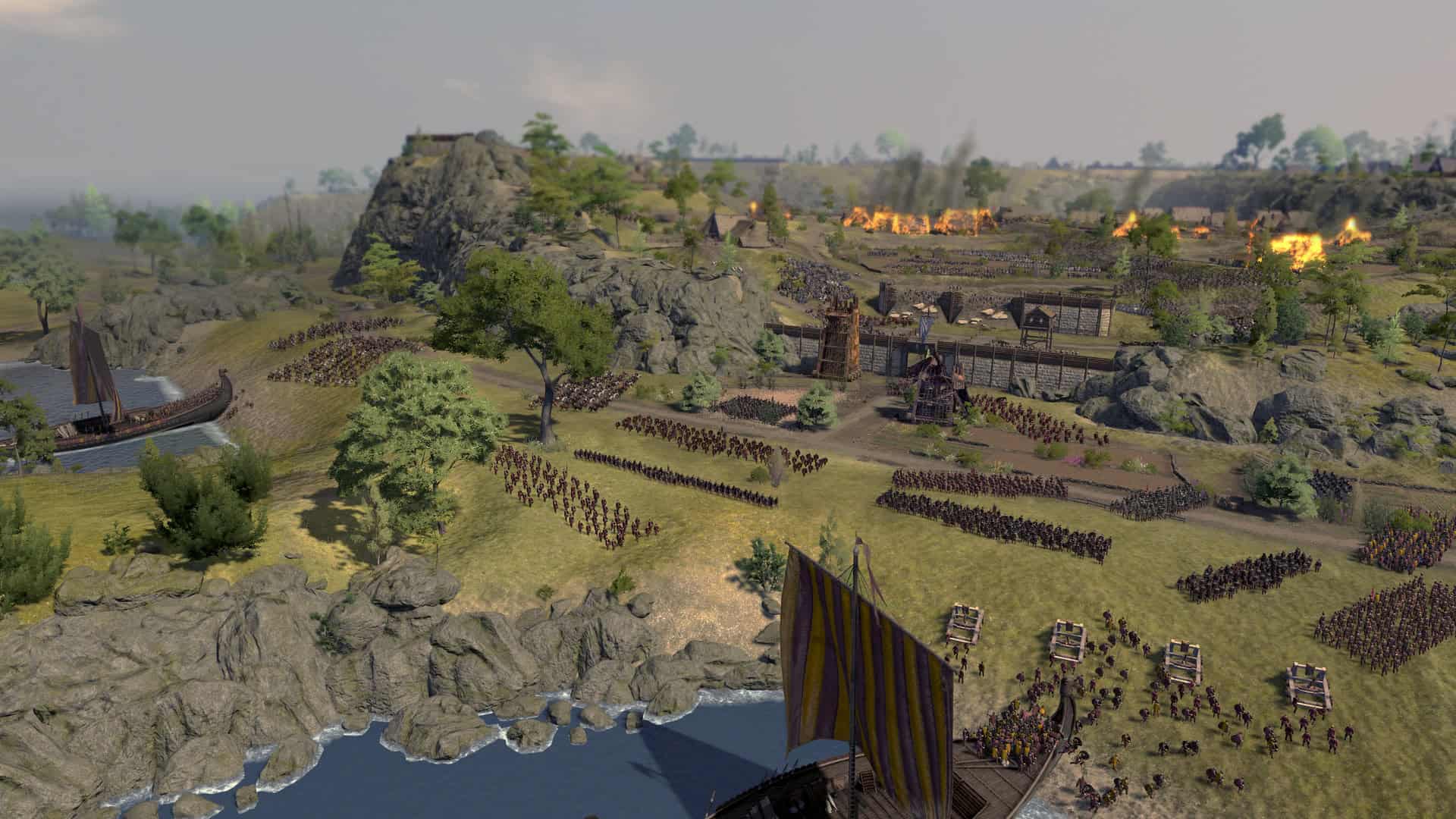 Large medieval battlefield with infantry, siege towers, and burning buildings near a rocky coast and a sailing ship